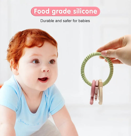 A hand holding a green Tipi Tipi Tap 4 in 1 silicone baby teether ring set in front of a baby with text about food-grade silicone.