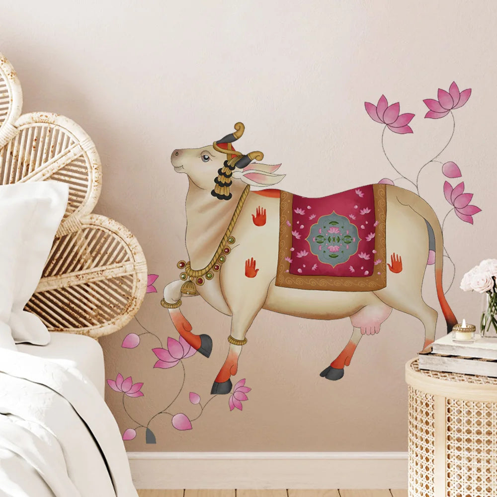 Tipi Tipi Tap Pichwai Art Gau Mata with Lotus Wall Sticker featuring a decorated cream-colored cow with pink lotus flowers, shown applied to a beige wall in a styled bedroom setting next to a woven headboard and bedside table.
