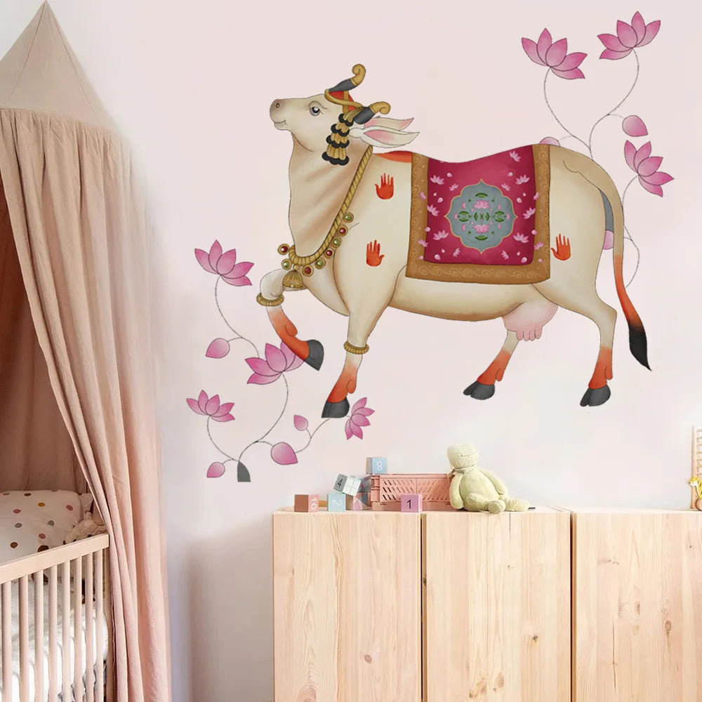 Tipi Tipi Tap Pichwai Art Gau Mata with Lotus Wall Sticker featuring a cream-colored cow with a pink and teal saddle, decorated with pink lotus flowers, shown applied to a light pink wall in a nursery setting above a wooden cabinet.