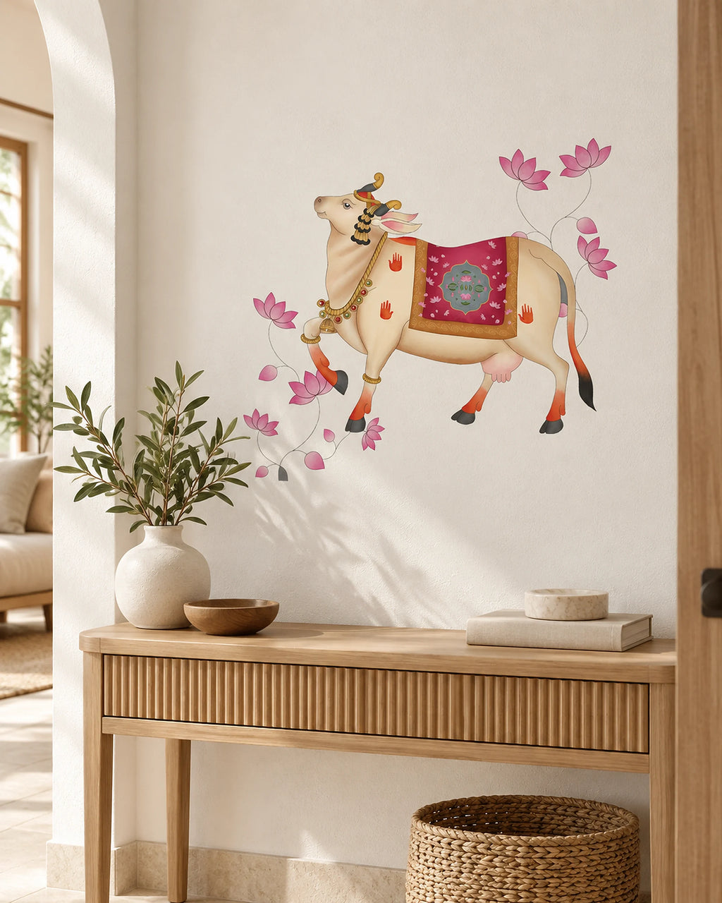 Tipi Tipi Tap Pichwai Art Gau Mata with Lotus Wall Sticker featuring a decorated cow with pink lotus flowers, shown applied to a white wall in a styled home interior above a wooden console table.