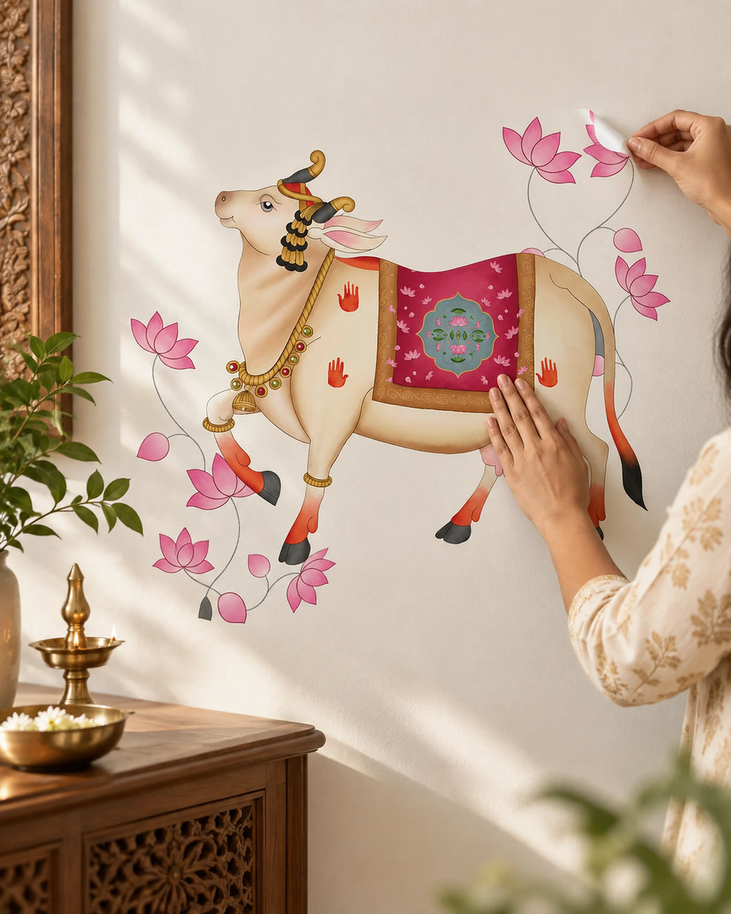 A person is applying Tipi Tipi Tap Pichwai Art Gau Mata with Lotus Wall Sticker featuring a decorated cow with pink lotus flowers on a white wall in a styled room setting with a wooden table and plants.