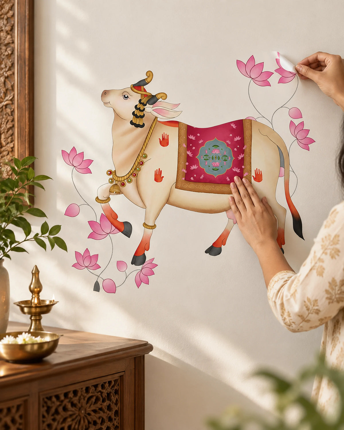 A person is applying Tipi Tipi Tap Pichwai Art Gau Mata with Lotus Wall Sticker featuring a decorated cow with pink lotus flowers on a white wall in a styled room setting with a wooden table and plants.