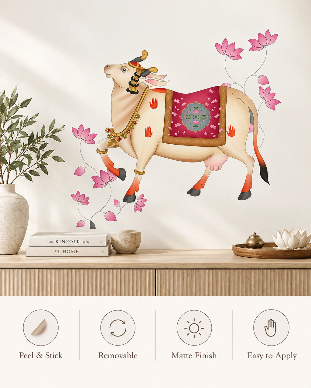 Tipi Tipi Tap Pichwai Art Gau Mata with Lotus Wall Sticker featuring a traditional Indian cow with pink lotus flowers, shown applied to a wall above a wooden console table with books and decor. Four feature icons at the bottom highlight peel-and-stick, removable, matte finish, and easy-to Apply qualities.