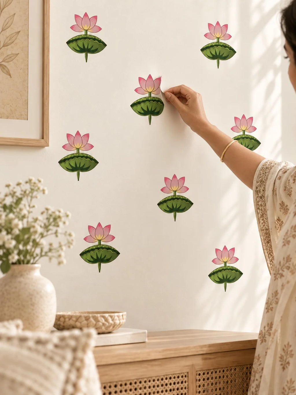 A person in a cream floral dress is shown from the side, placing a Tipi Tipi Tap Pink Lotus Flower Wall Stickers on a warm off-white wall. Other matching decals are already applied to the wall.