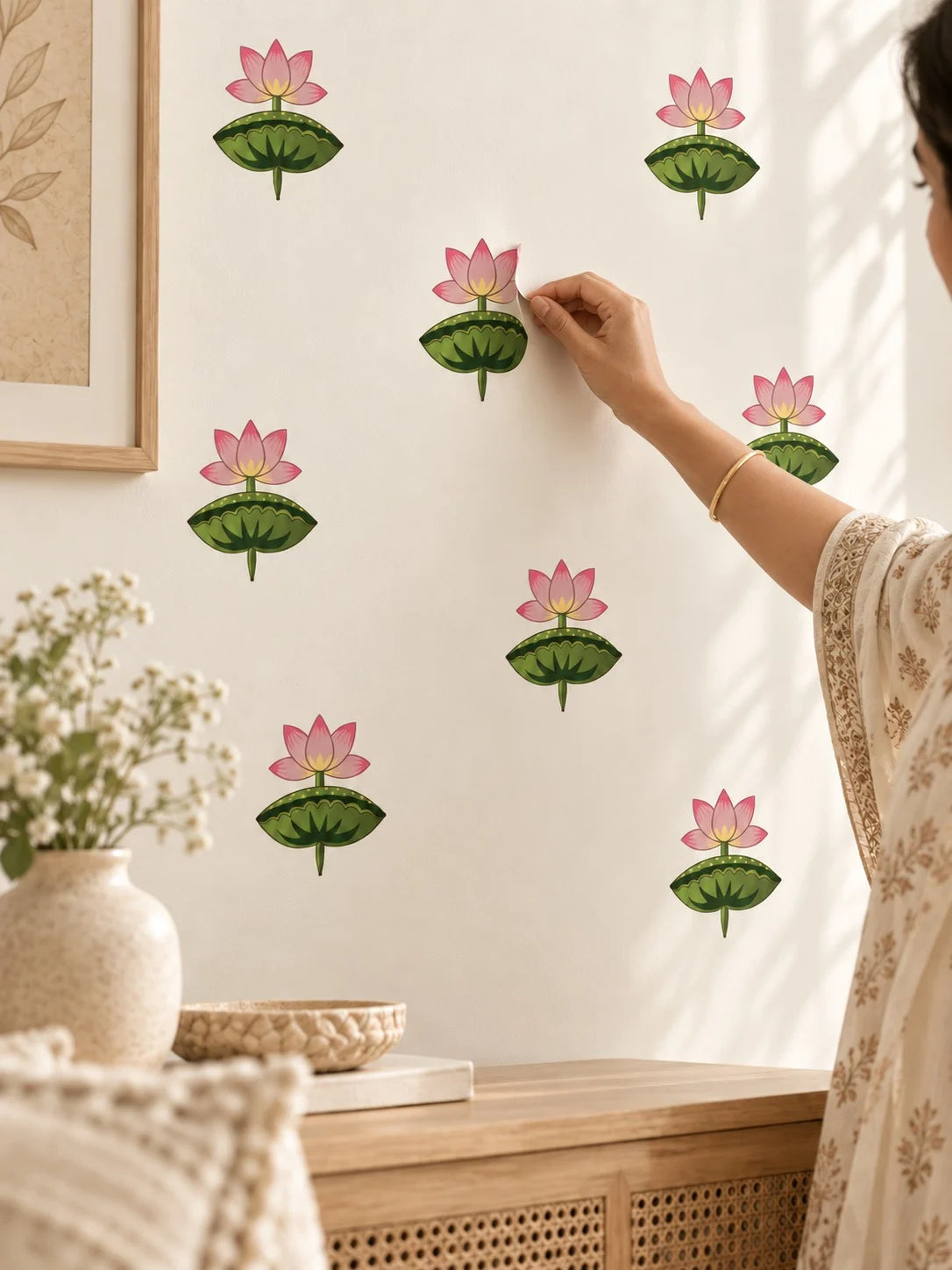 A person in a cream floral dress is shown from the side, placing a Tipi Tipi Tap Pink Lotus Flower Wall Stickers on a warm off-white wall. Other matching decals are already applied to the wall.