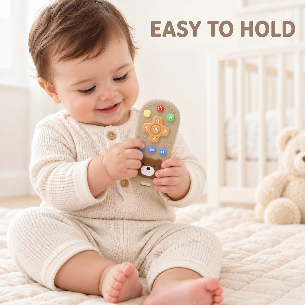 A baby sitting on a quilted cream bed holds a plush Tipi Tipi Tap Silicone Baby Remote Teether. The baby is wearing a cream waffle-knit romper, and a beige teddy bear is visible in the background.