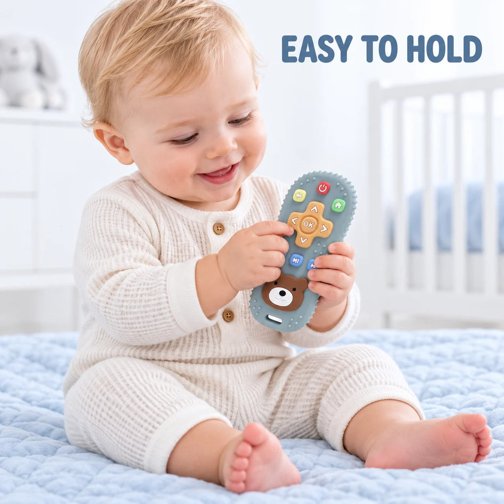 A baby sitting on a light blue quilted bed holds a blue Tipi Tipi Tap Silicone Baby Remote Teether with colorful buttons and a bear face. The baby is wearing a cream waffle-knit romper.