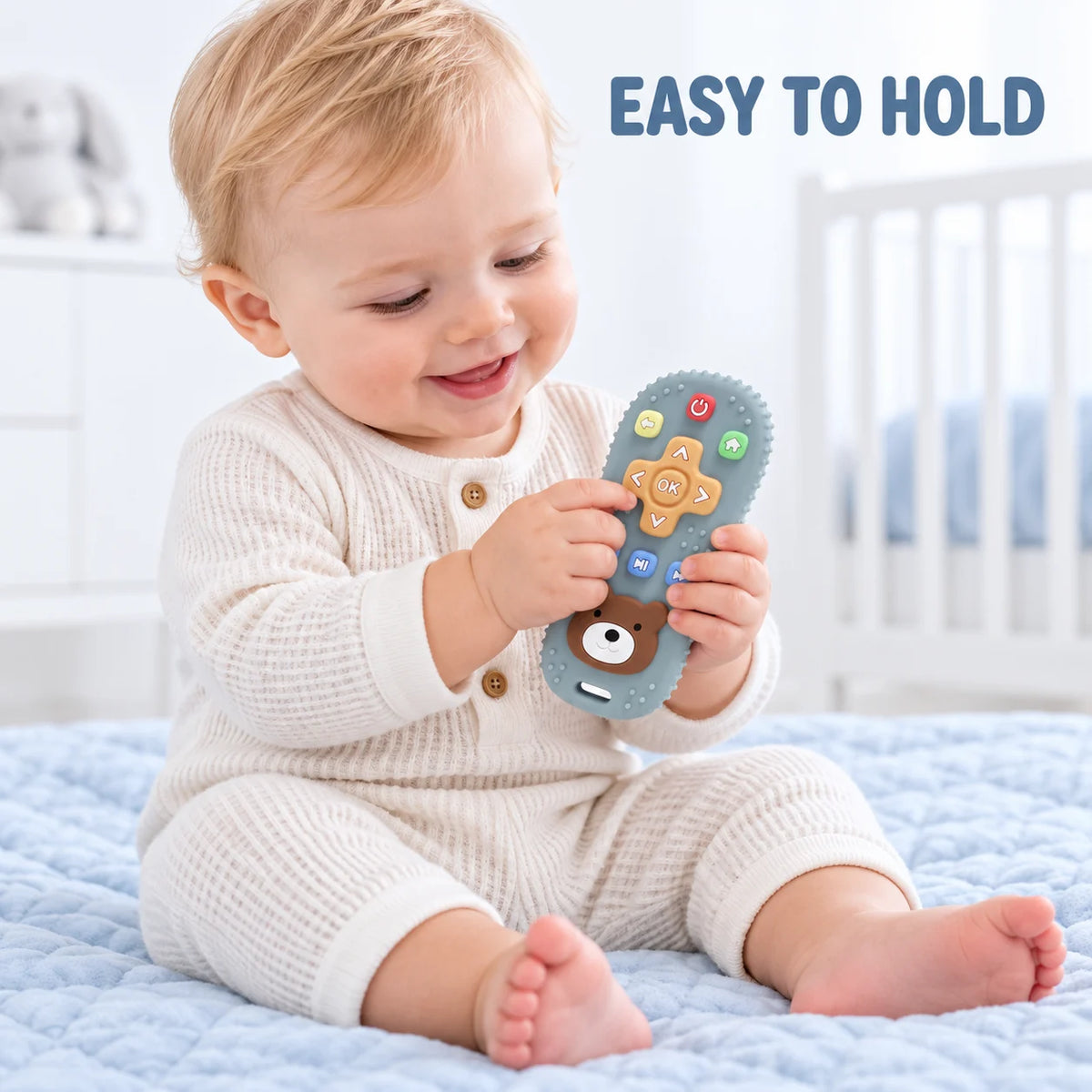 A baby sitting on a light blue quilted bed holds a blue Tipi Tipi Tap Silicone Baby Remote Teether with colorful buttons and a bear face. The baby is wearing a cream waffle-knit romper.