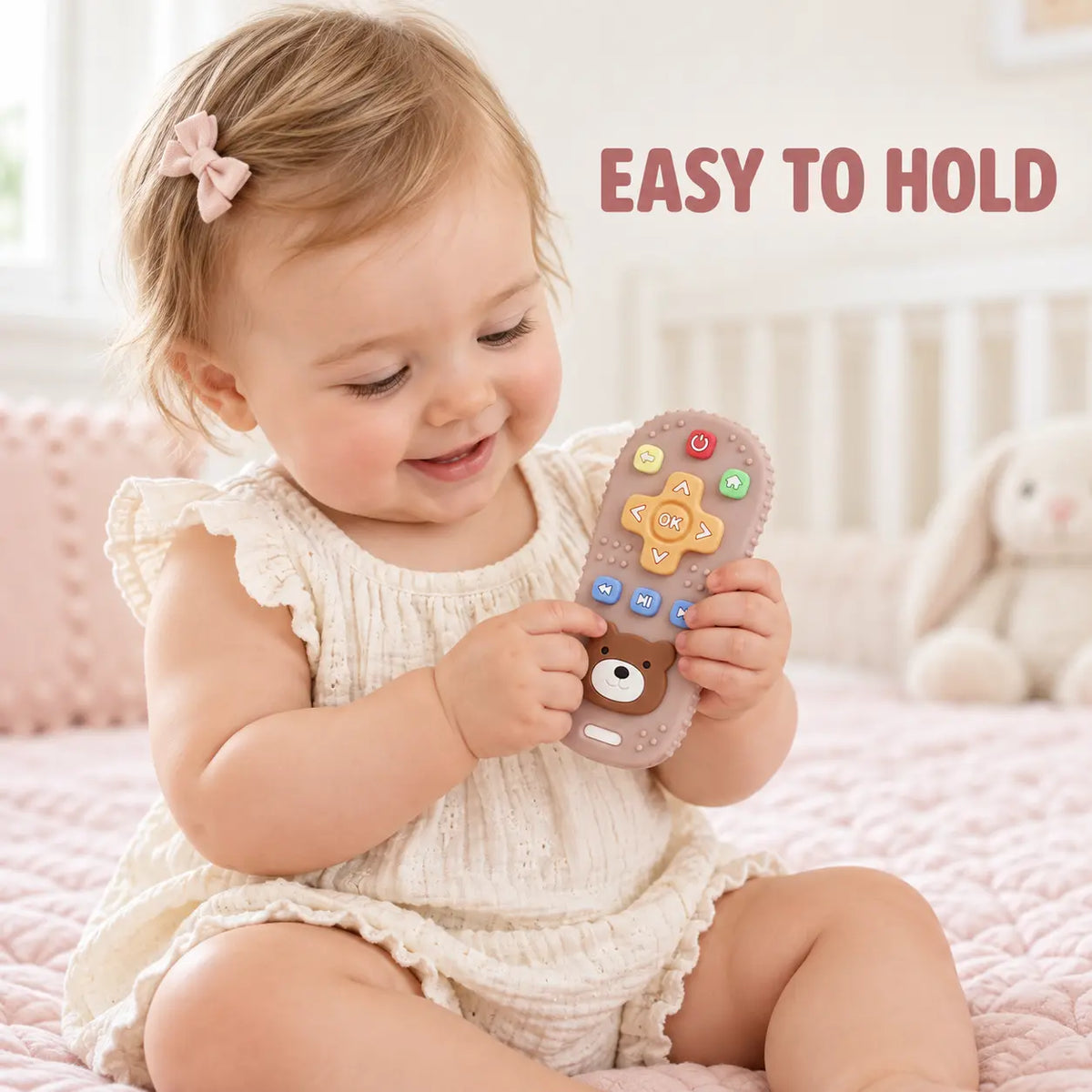 A baby girl in a cream-colored romper sits on a pink quilted bed, holding Tipi Tipi Tap Silicone Baby Remote Teether Toy. The teether is light brown with colorful buttons and a cartoon bear face.
