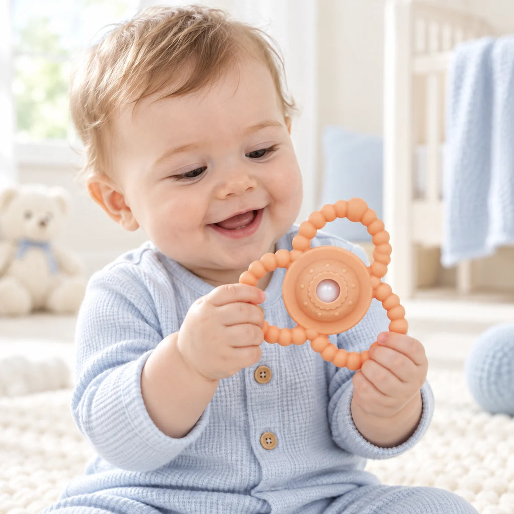 A baby wearing a light blue waffle-knit onesie sits on a cream rug, smiling and holding an orange Tipi Tipi Tap Triangular Rattle Teether.