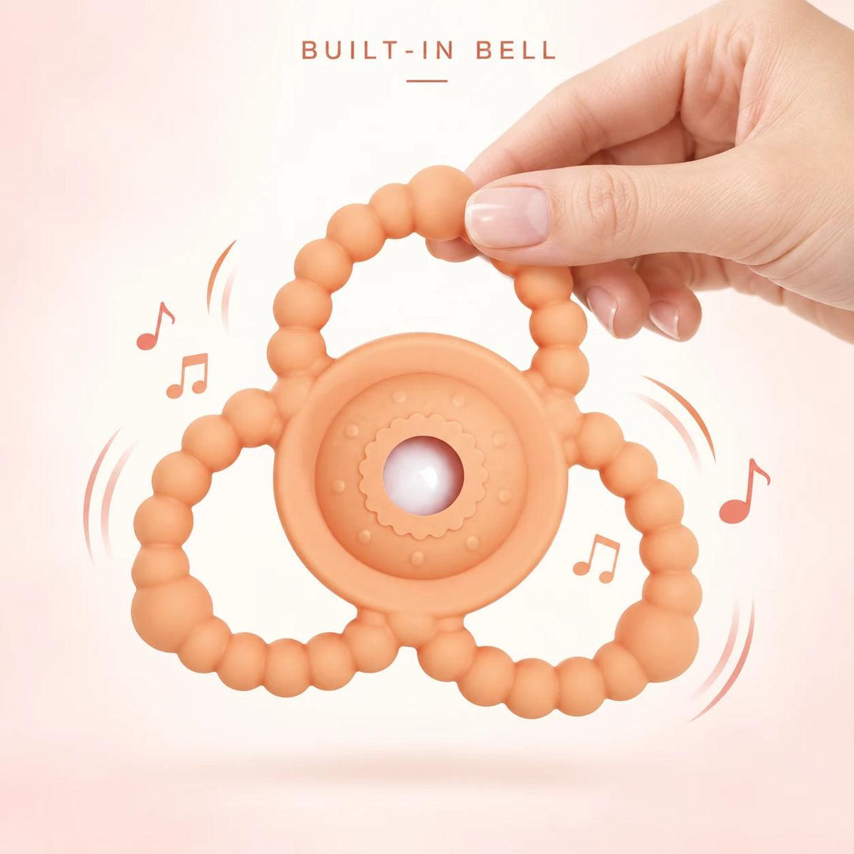 An orange silicone Tipi Tipi Tap Triangular Rattle Teether, held by a hand. The teether features a central round textured piece with a small white bell in the middle, connected to a circular handle made of small orange beads.
