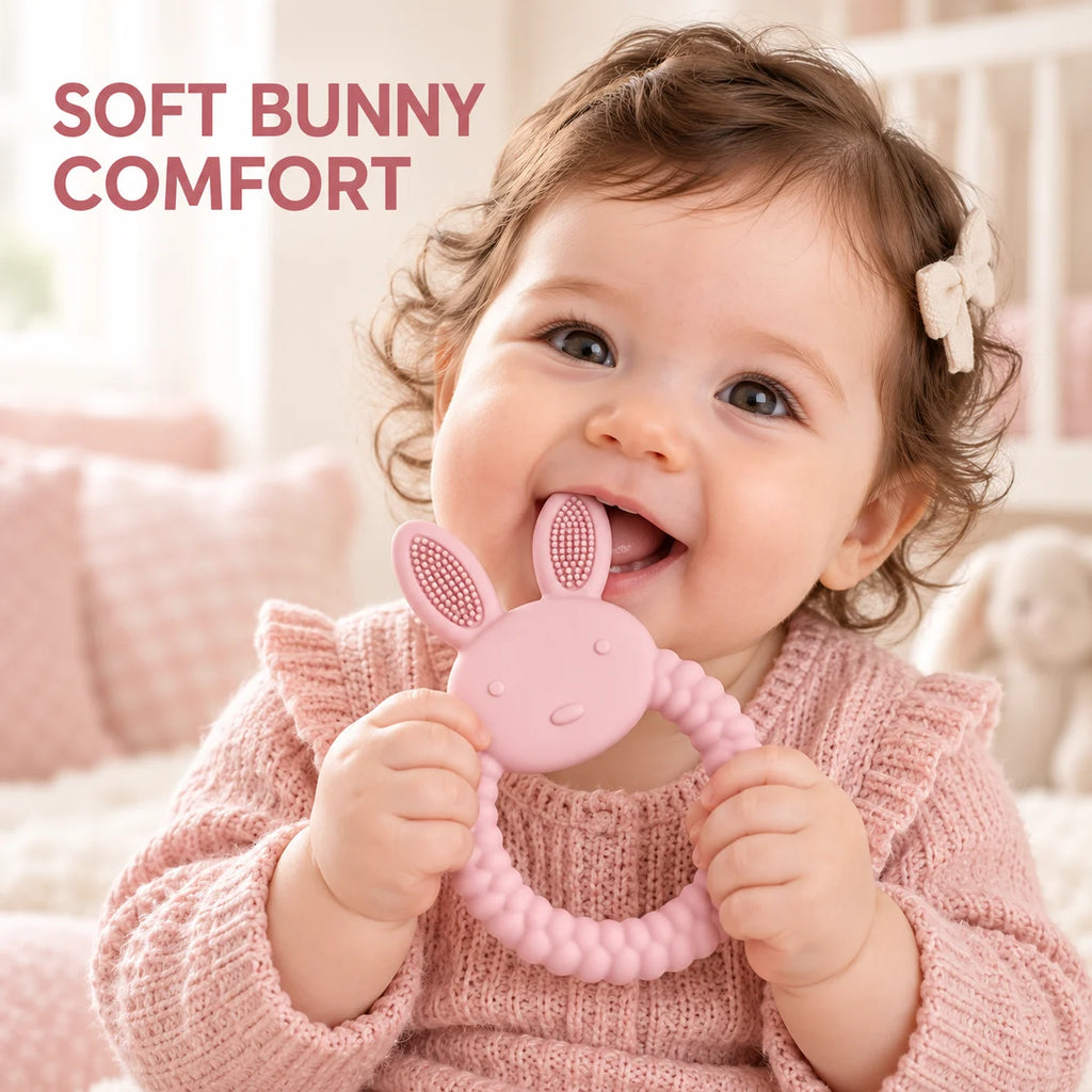 A baby wearing a pink knit sweater holds a pink Tipi Tipi Tap Rabbit Silicone Baby Teether in her mouth. The background is a soft, warm, blurred nursery setting with pink pillows, and the text 'SOFT BUNNY COMFORT' is displayed in the top left corner.
