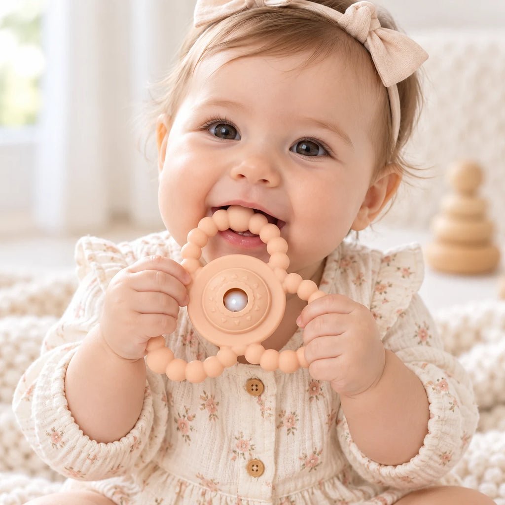A baby wearing a cream floral dress and a beige bow headband holds a peach-colored silicone Tipi Tipi Tap Triangular Rattle Teether in their mouth, set against a soft, warm, neutral background.