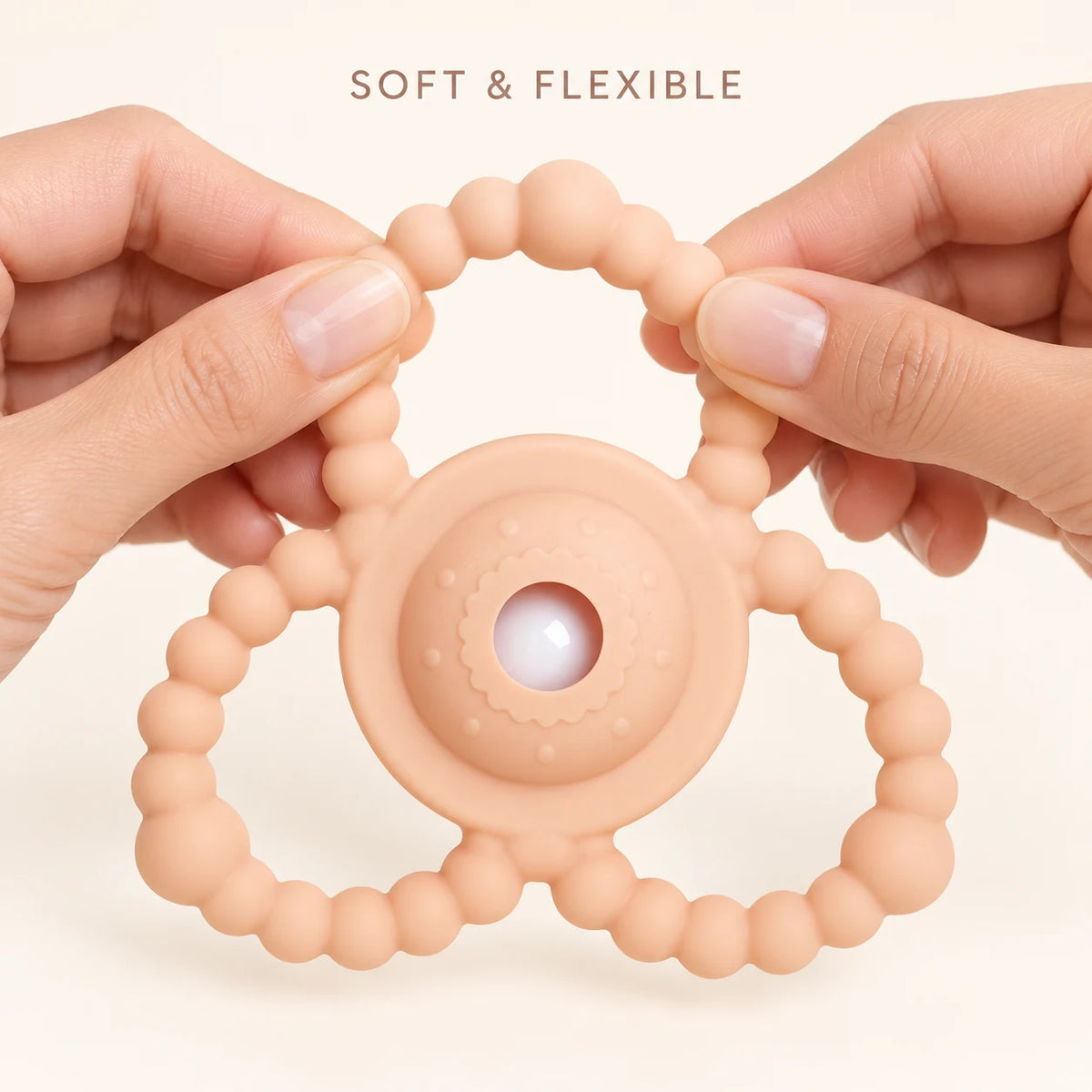A peach-colored silicone Tipi Tipi Tap Triangular Rattle baby teether with a beaded ring and central wavy texture, held by two hands against a light cream background with the text 'SOFT & FLEXIBLE' at the top.