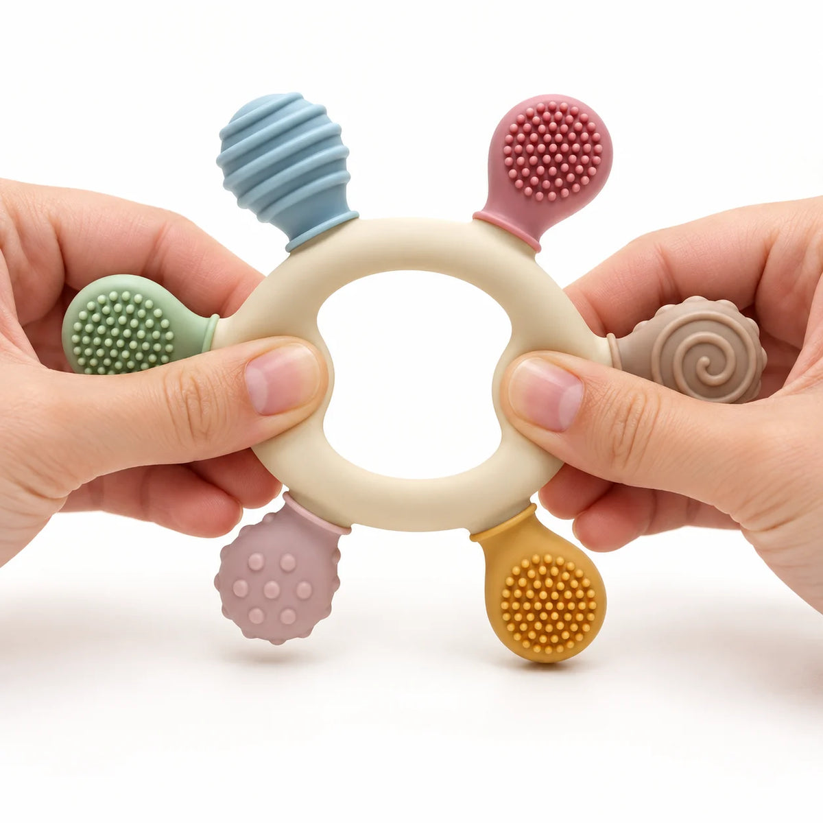 Tipi Tipi Tap Rainbow Ring Silicone Baby Teether held by two hands against a white background. The cream-colored ring has six textured, colorful sensory tabs: a blue honeycomb, a pink bristle brush, a pink textured ball, a yellow bristle brush, and a beige spiral.