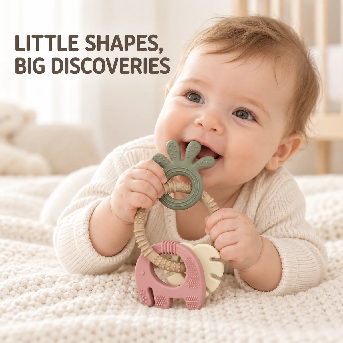 A baby lying on a cream knit blanket holds a Tipi Tipi Tap 4-in-1 Silicone Baby Teether Ring Set with a sage green leaf-shaped ring and a pink elephant-shaped piece attached to a beige rope. The text 'LITTLE SHAPES, BIG DISCOVERIES' appears in the upper left.