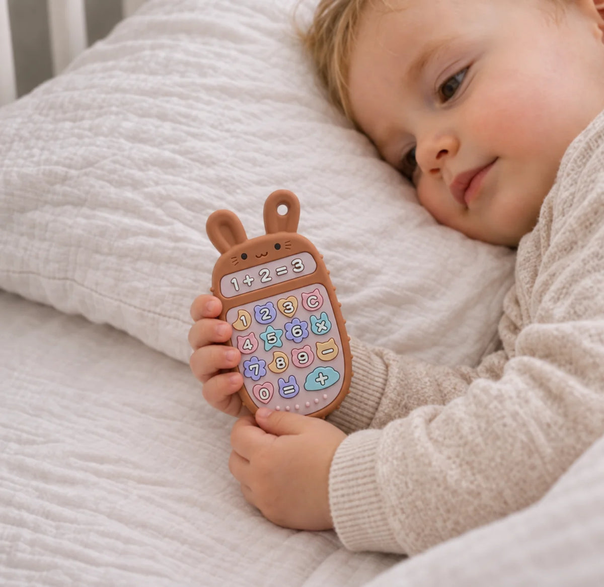 A baby lying on a white quilted bed holds a brown Tipi Tipi Tap Bunny Calculator Silicone Baby Teether with colorful buttons.