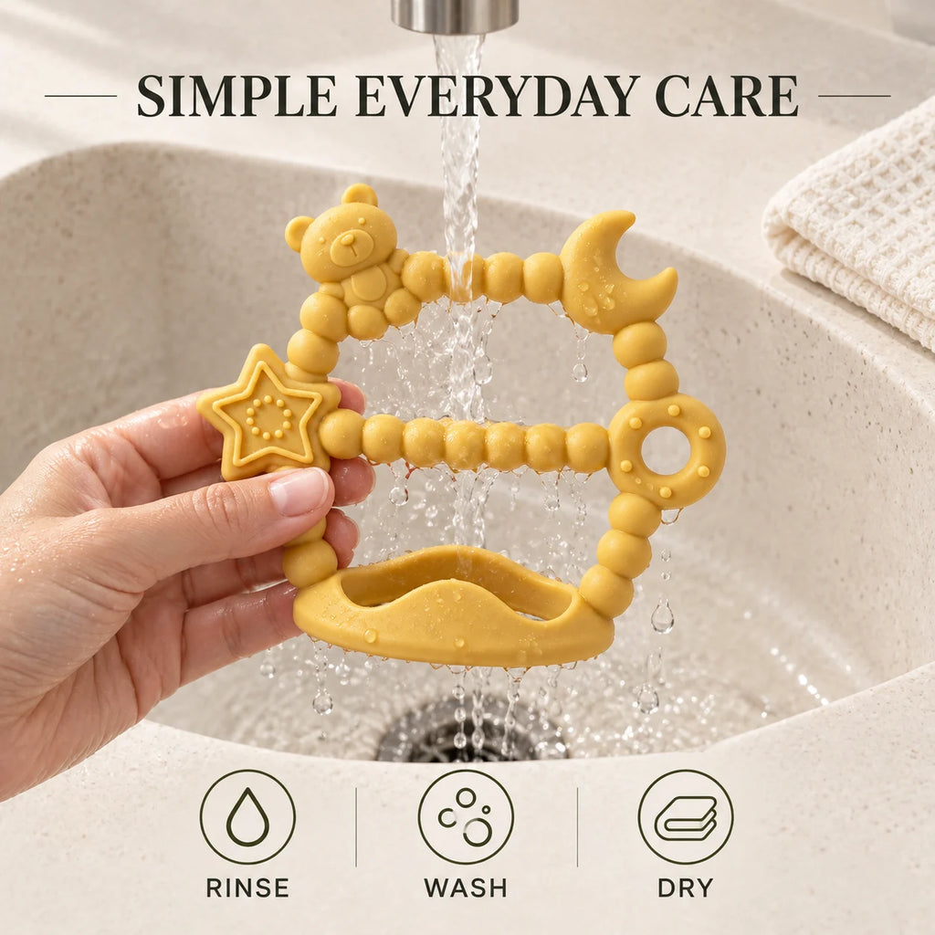 A hand holds a yellow Tipi Tipi Tap Silicone Baby Wrist Teether (Moon & Star) under running water in a sink. The teether has a bear head, moon, star, and curved arrow design.